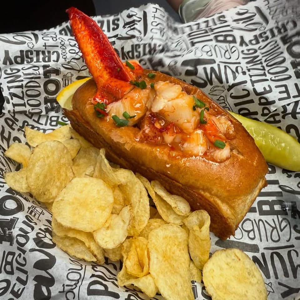 The Hungry Captain's lobster roll with a claw, chips, lemon, and pickle on branded paper.