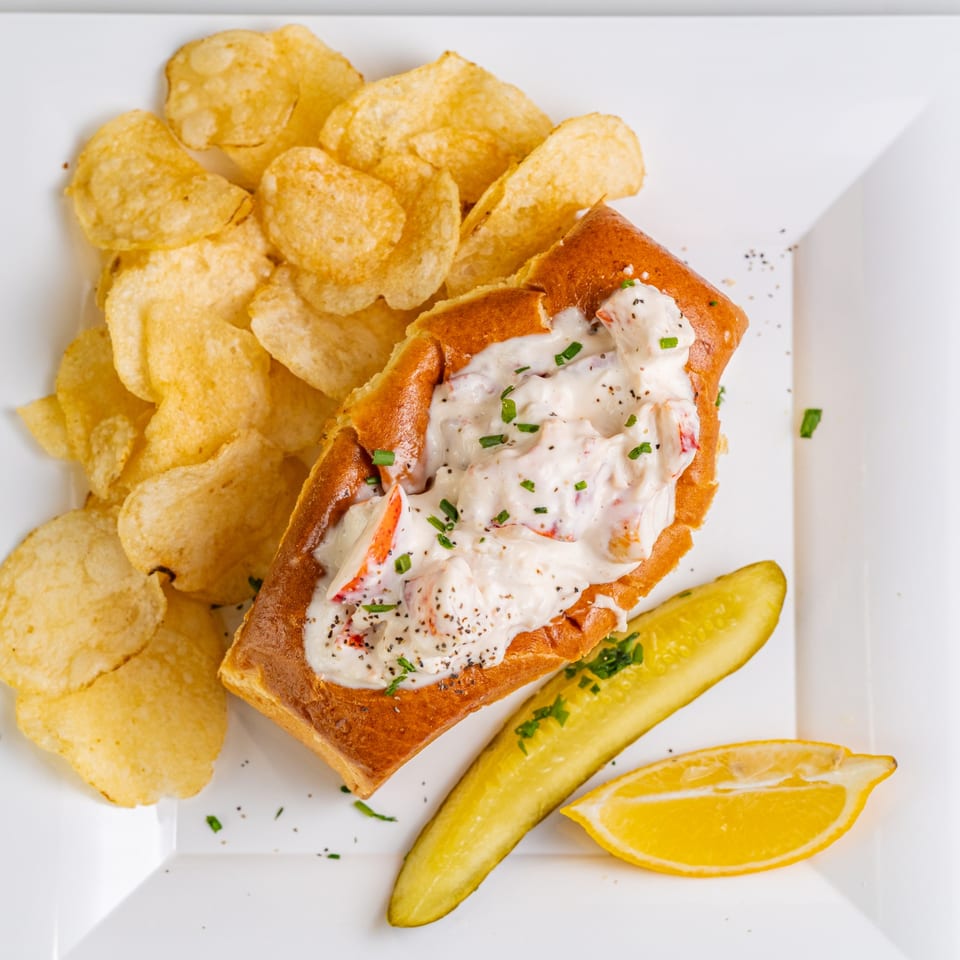 The Hungry Captain's lobster roll with creamy filling, chips, a pickle spear, and lemon wedge on a white plate.