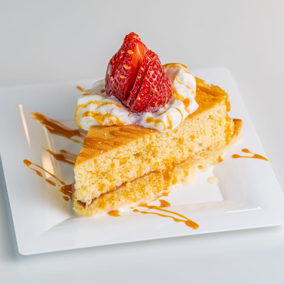 Slice of golden cake with whipped cream, caramel drizzle, and a sliced strawberry on a white plate.