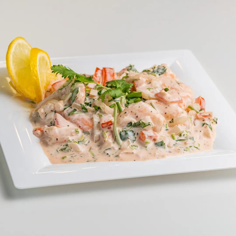 Creamy shrimp ceviche with red peppers, cilantro, and lemon wedges on a white square plate.